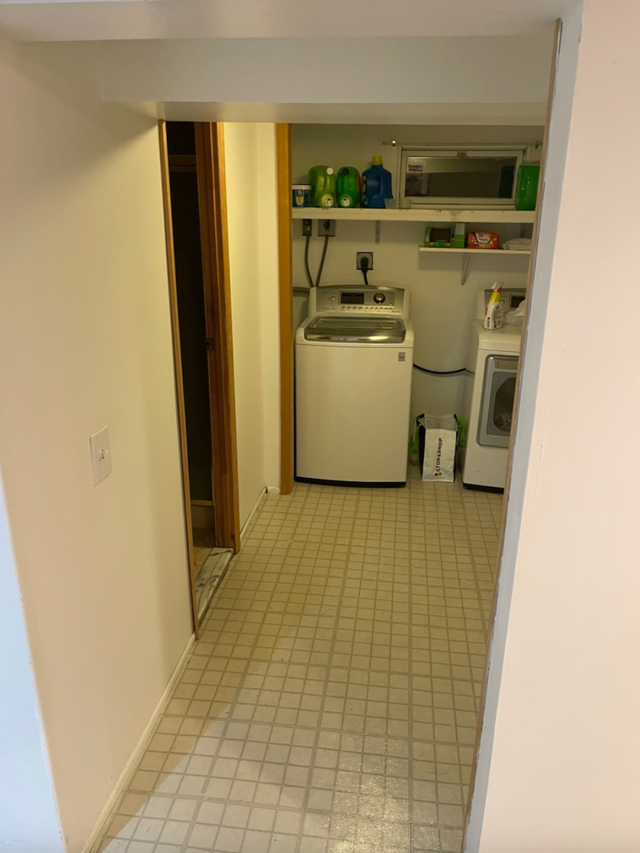 Laundry room with a washing machine, dryer, and shelves. Light-colored walls, tiled floor, and a doorway.