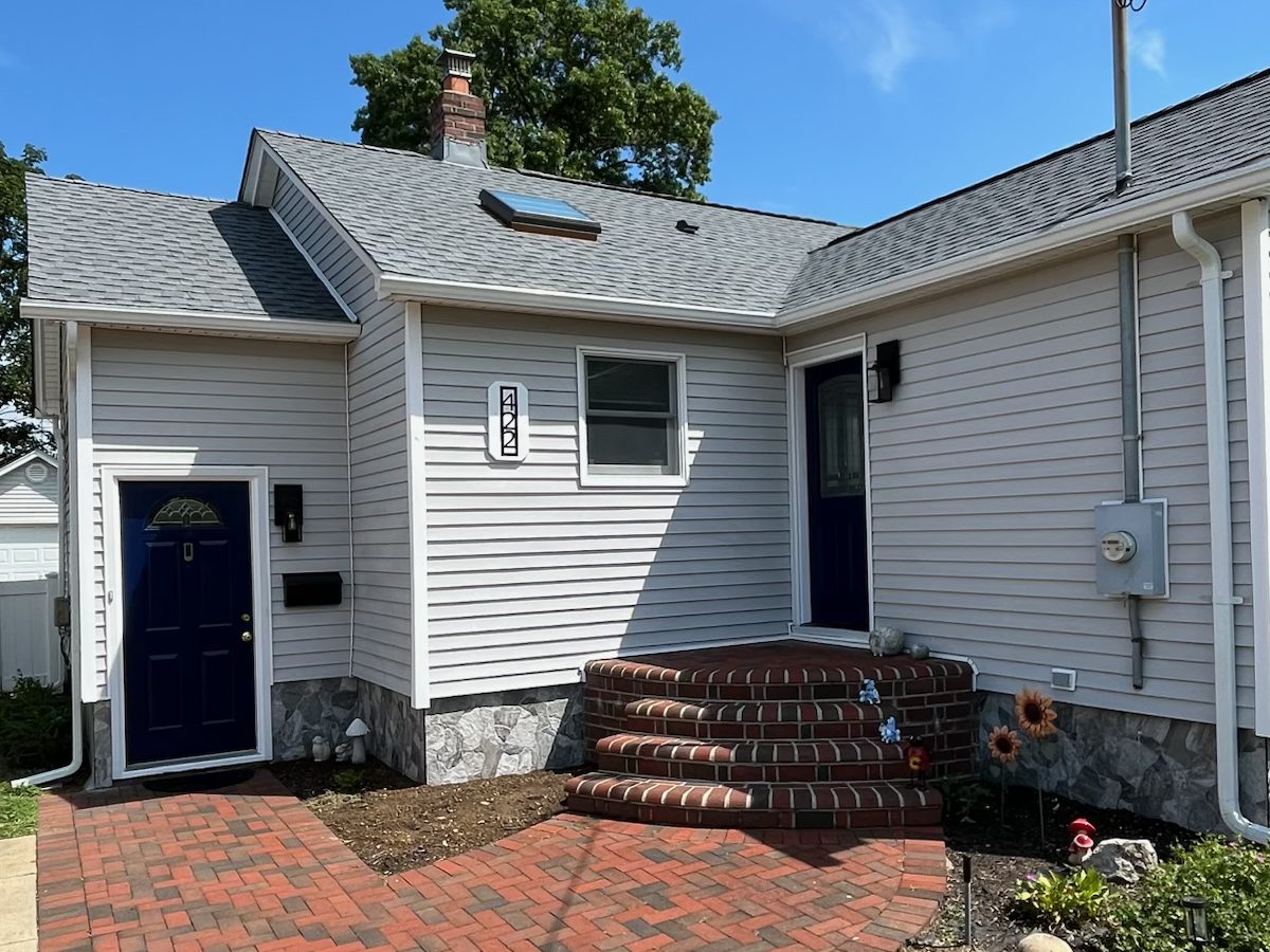 Finished L.I. Taylor job in Bellmore, NY: new grey architectural shingle roof on a cape with white siding, red brick steps and a navy front door, July 2026
