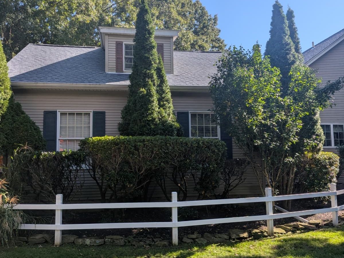 Side of the Southold cape with its new light grey shingle roof and a side dormer, behind evergreens and a white rail fence
